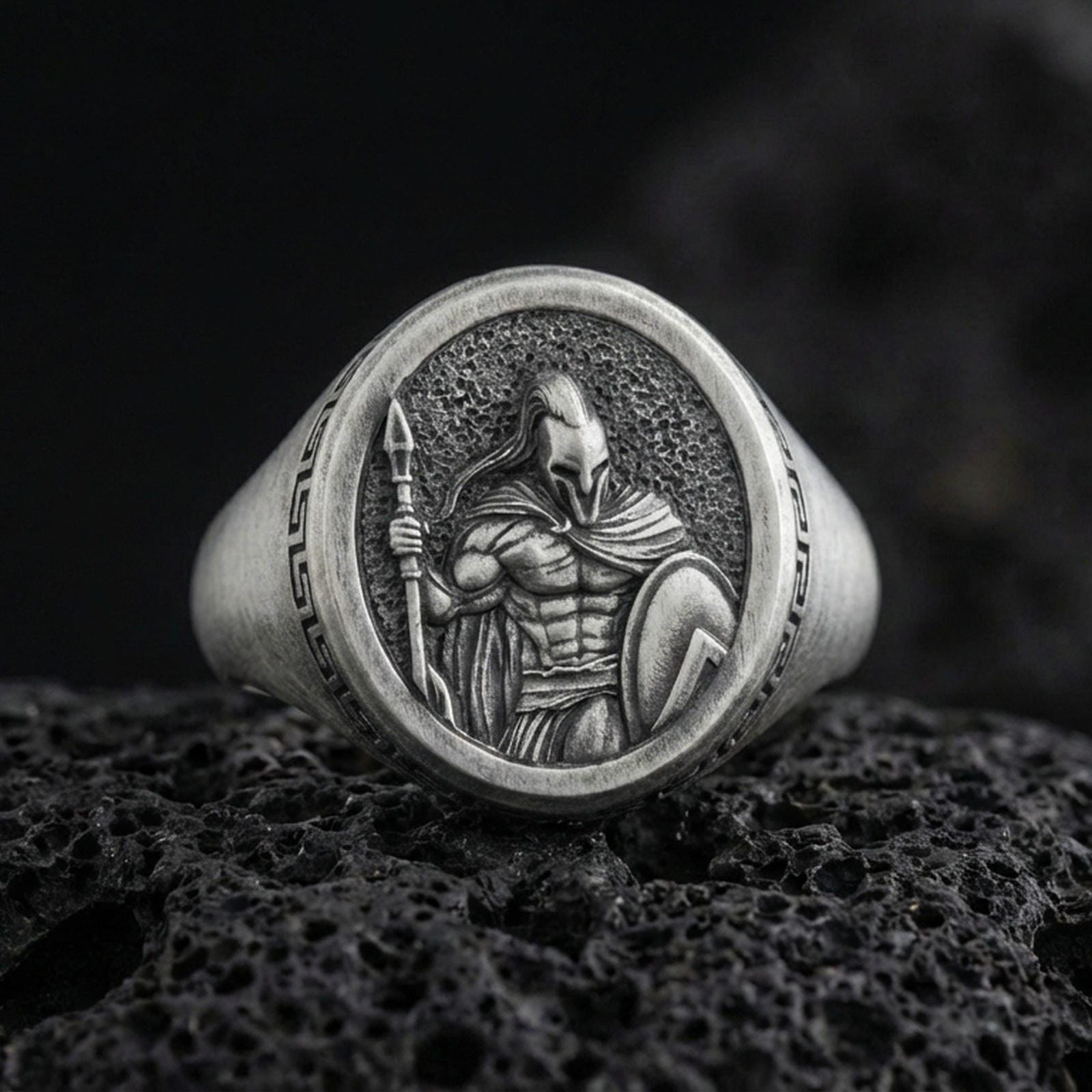 Oxidized sterling silver Spartan warrior signet ring with spear and shield relief, photographed on black volcanic rock in a dark macro studio