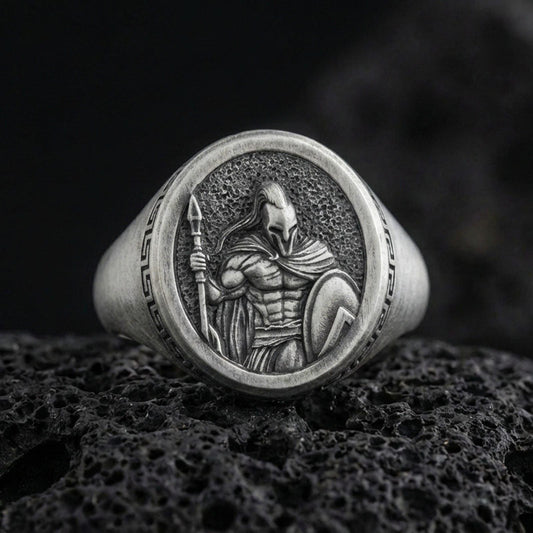 Oxidized sterling silver Spartan warrior signet ring with spear and shield relief, photographed on black volcanic rock in a dark macro studio