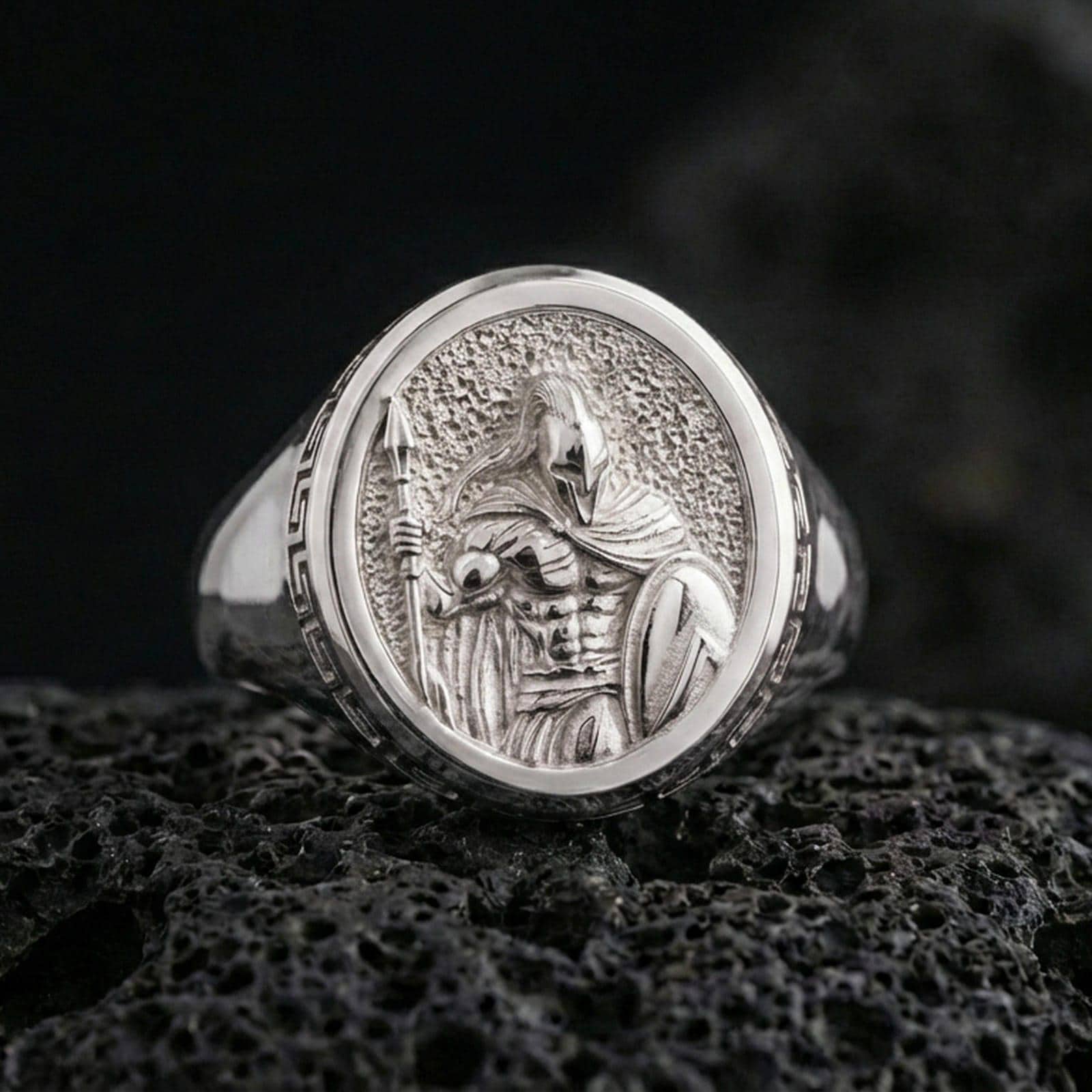 Polished silver Greek warrior signet ring with high-relief helmeted figure and crisp highlights, macro close-up on textured volcanic stone