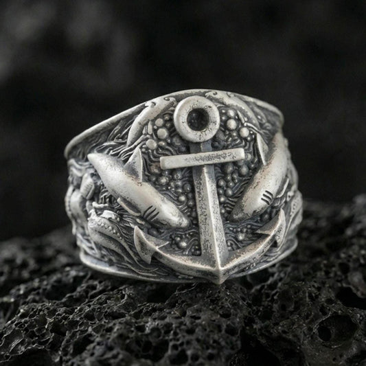 Oxidized sterling silver anchor ring with surrounding shark motifs and ocean-texture details, macro product photo on black volcanic rock