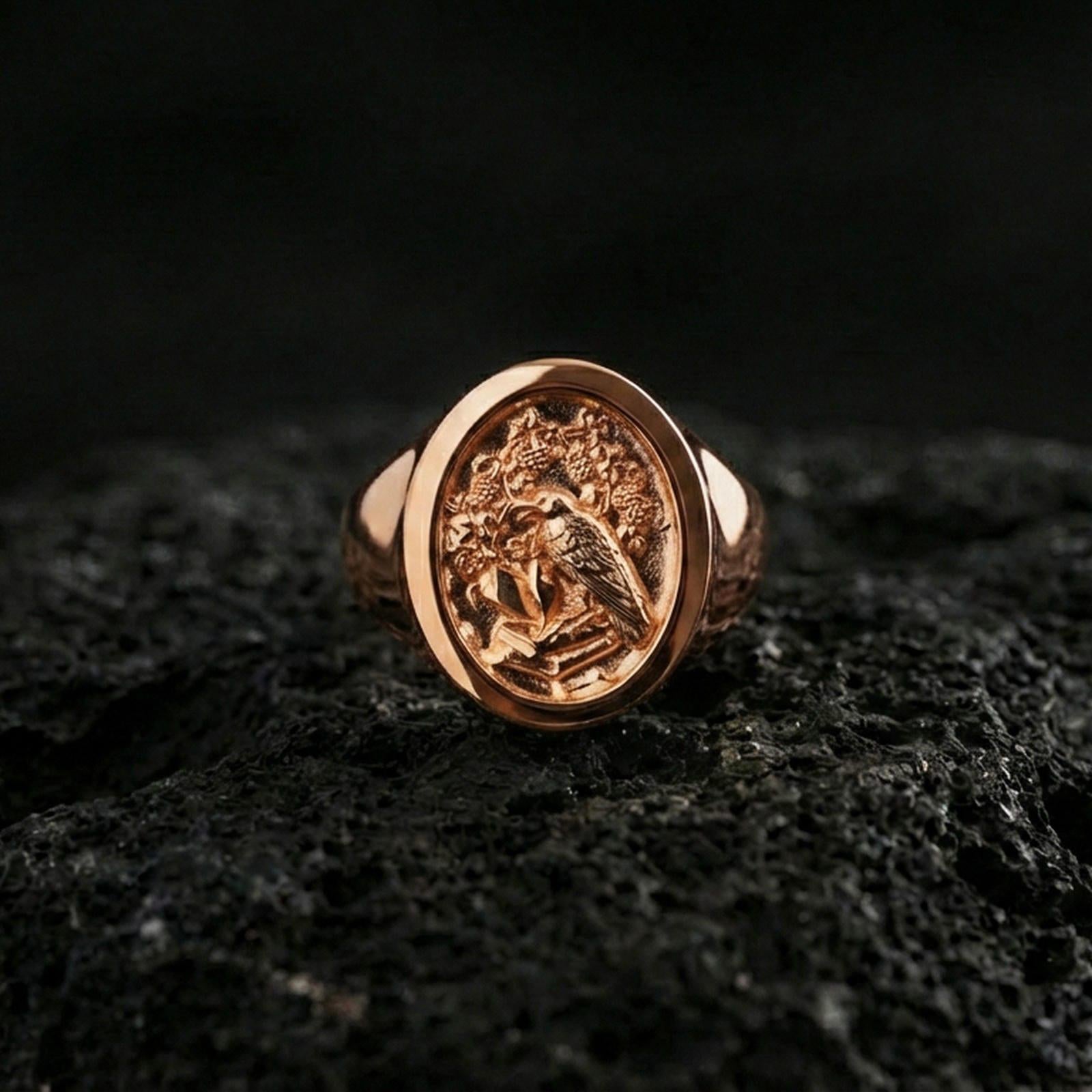 Rose gold plated raven signet ring with ornate emblem engraving and vintage-style relief, macro product shot on volcanic rock surface