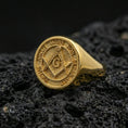 Load image into Gallery viewer, Gold plated Masonic signet ring with square and compass “G” symbol and Latin inscription, product-only macro shot on black volcanic rock
