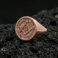Load image into Gallery viewer, Rose gold plated Freemason signet ring with engraved square and compass emblem and motto ring border, macro image on volcanic rock
