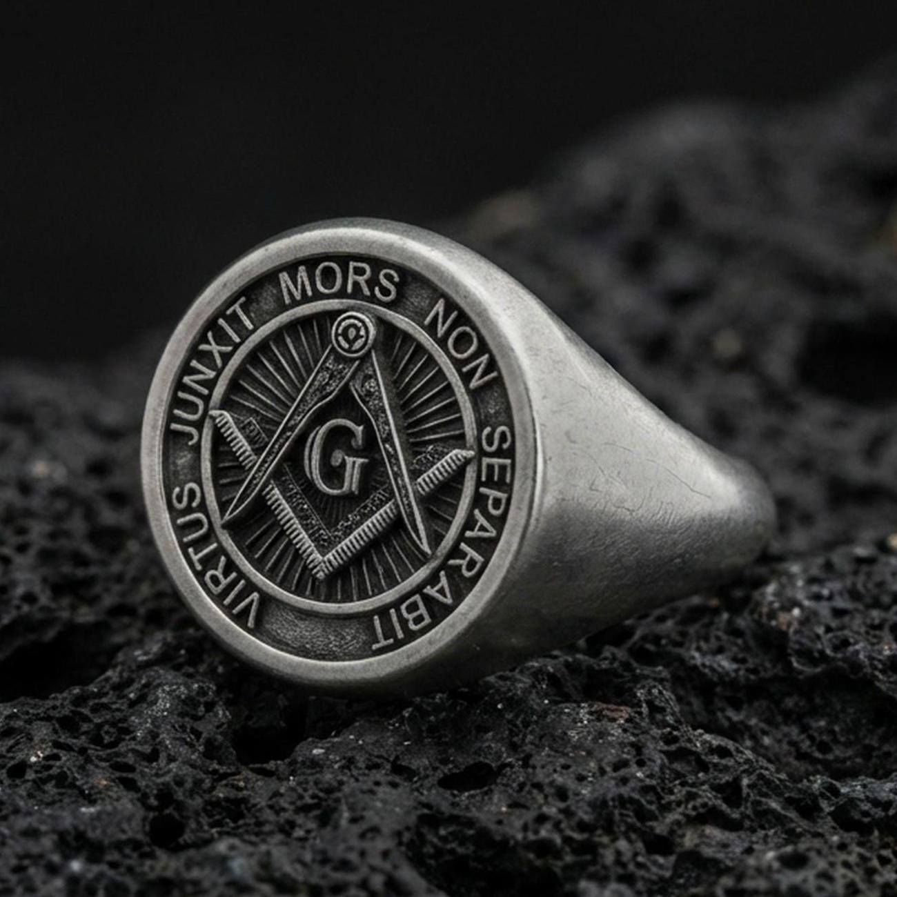 Oxidized sterling silver Masonic signet ring with square and compass “G” emblem and Latin motto, macro photo on black volcanic rock