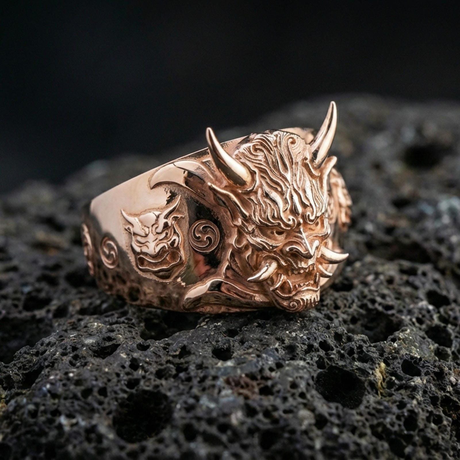 Rose gold plated Japanese demon mask ring with detailed Oni facial carving and side symbol engraving, macro image on volcanic rock