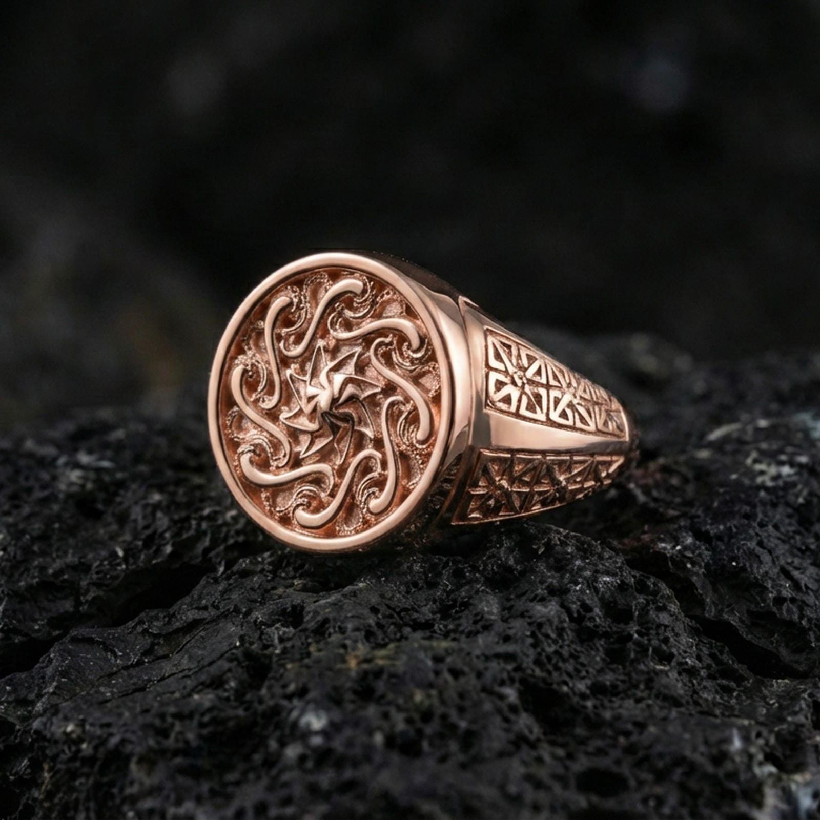 Rose gold plated round spiral signet ring with ornate swirling face design and patterned side panels, macro image on volcanic rock