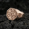 Load image into Gallery viewer, Rose gold plated octagonal signet ring with Celtic triskelion swirl design and side symbol detail, product-only macro shot on volcanic rock
