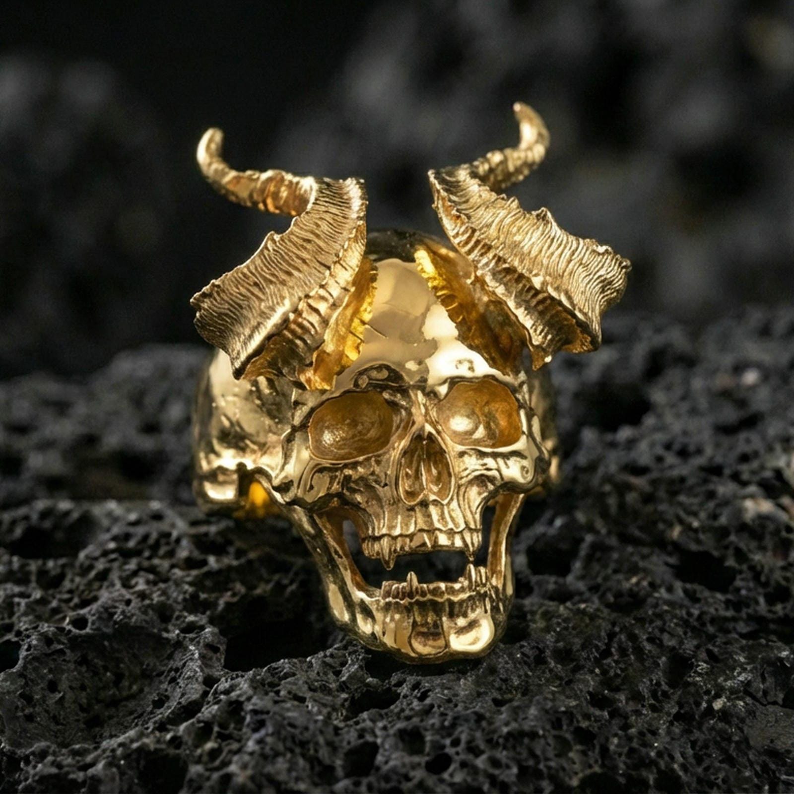 Gold plated demon skull ring with raised broken horns and detailed jawline, macro product-only image on black volcanic rock