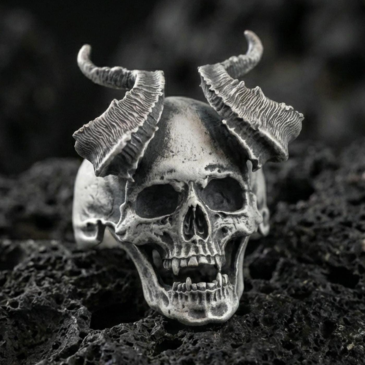 Oxidized sterling silver demon skull ring with cracked ram horns and hollow eye sockets, macro product photo on black volcanic rock