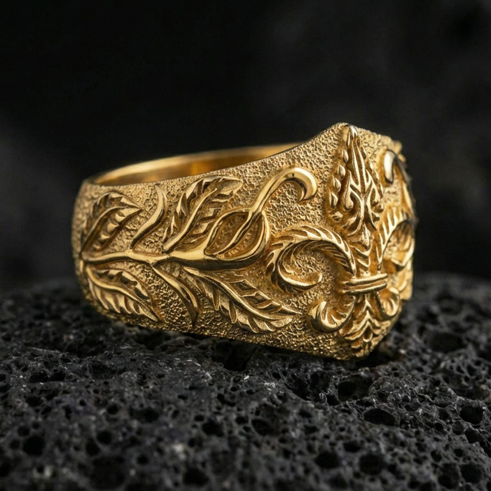 Gold plated fleur-de-lis signet ring with vine engraving and bold crest relief, macro product-only image on black volcanic rock