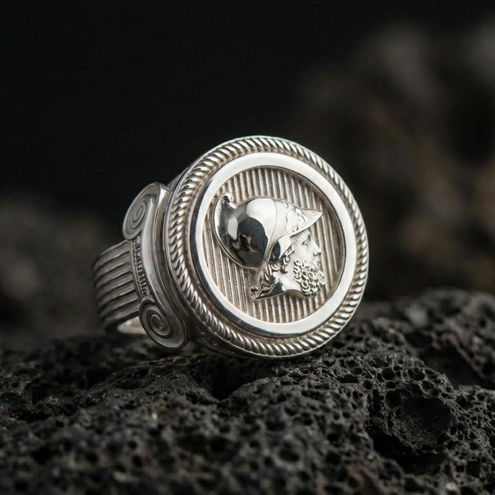 Polished silver coin-style ring featuring a helmeted warrior head and striped background texture, close-up shot on volcanic stone