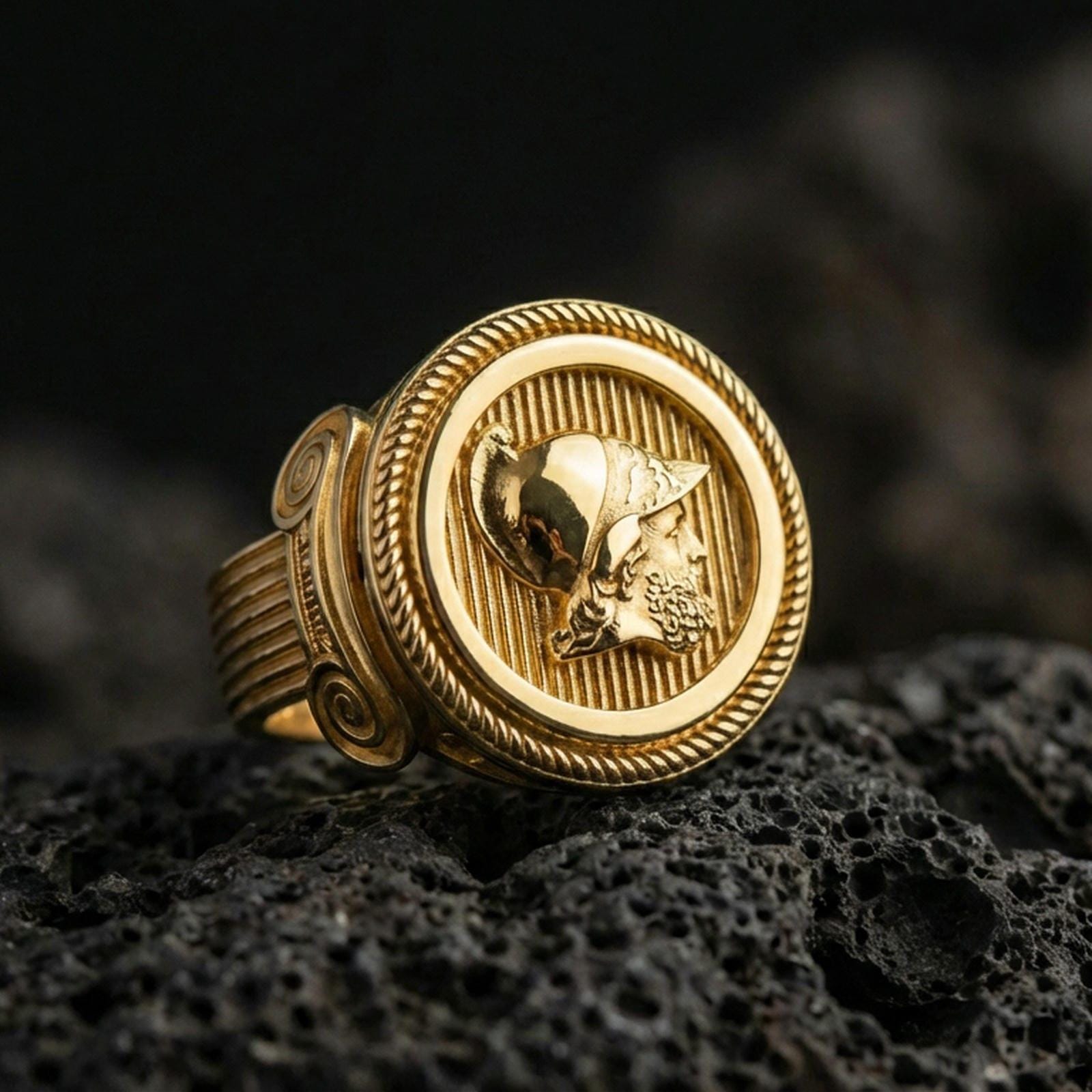 Gold plated Greek coin signet ring with raised warrior profile and braided rope border, macro product-only image on black volcanic rock