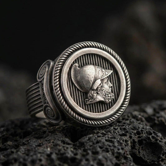 Oxidized sterling silver Greek coin ring with warrior profile relief and rope-edge frame, macro product photo on black volcanic rock