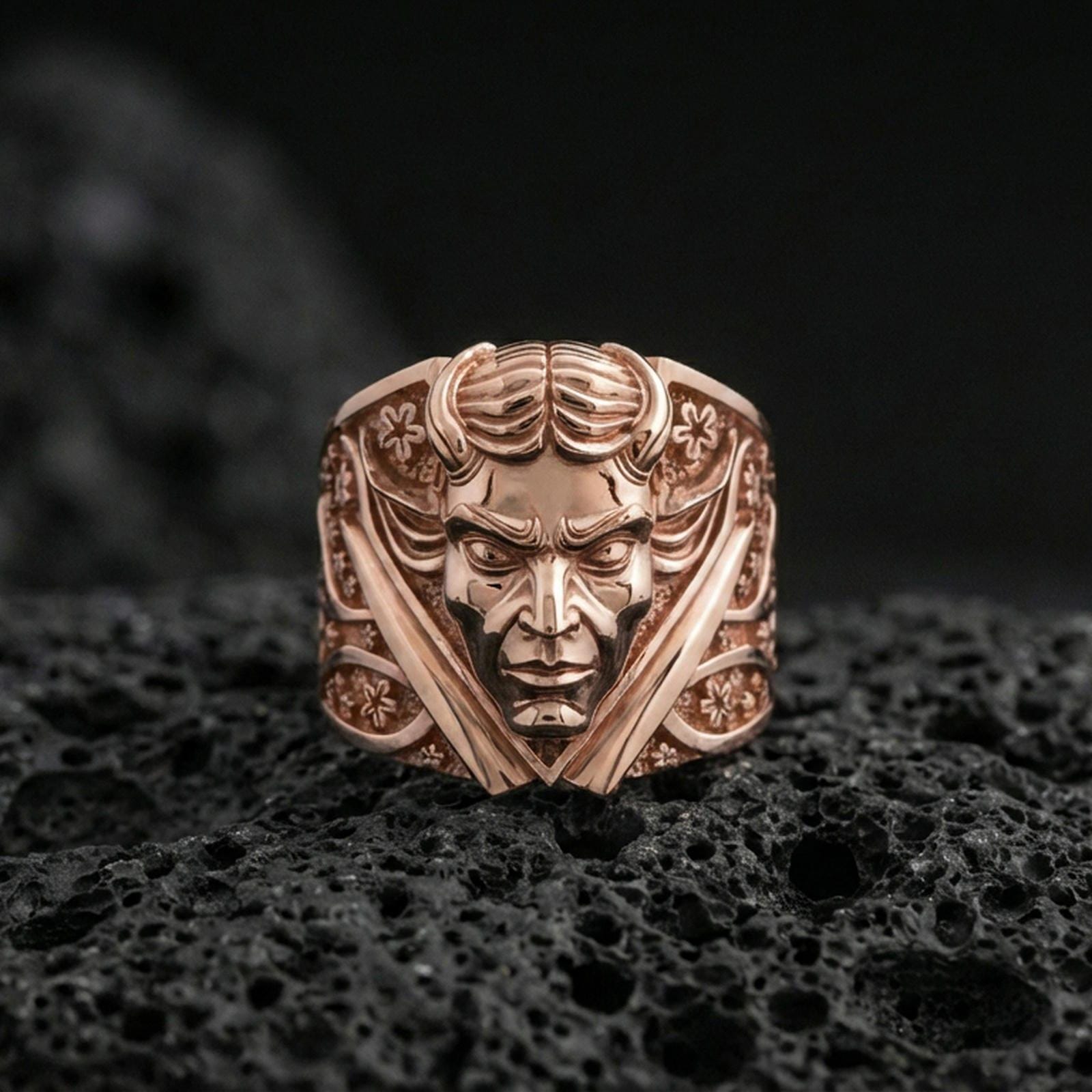 Rose gold plated demon face signet ring with crossed blades and decorative patterning, close-up photo on volcanic rock