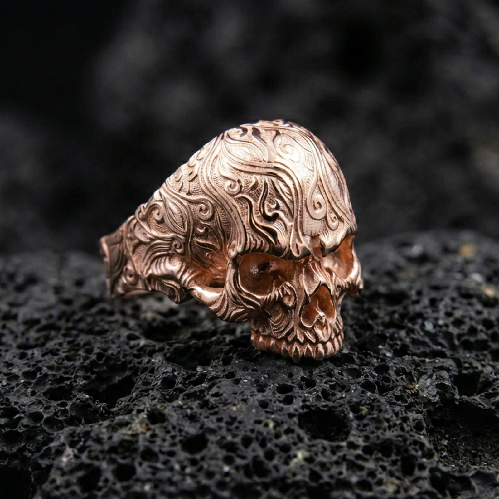 Rose gold plated gothic skull ring with intricate engraved scroll motifs and sculpted jaw detail, close-up on volcanic rock