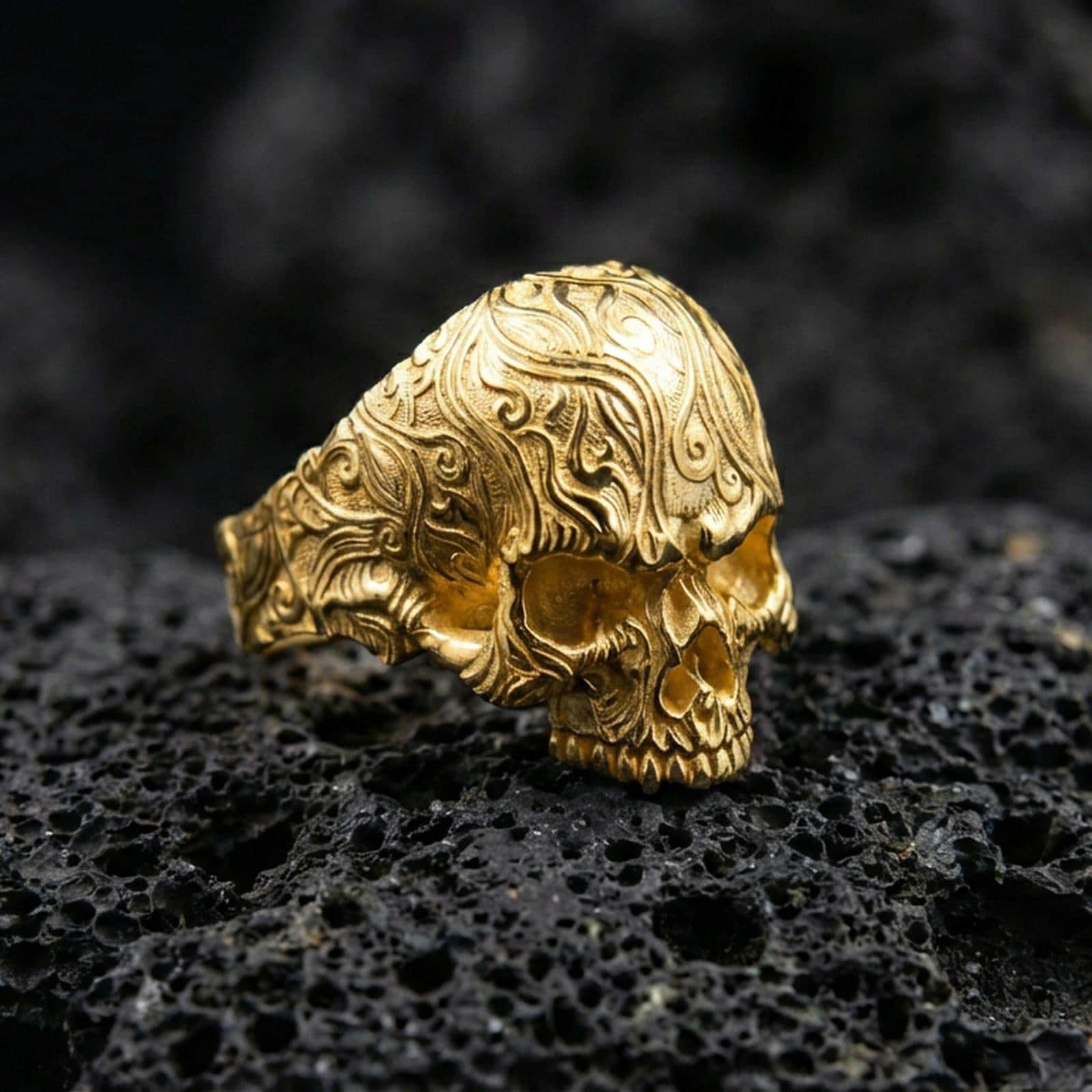 Gold plated skull ring with detailed baroque scrollwork pattern and high-relief engraving, product-only macro image on black volcanic rock