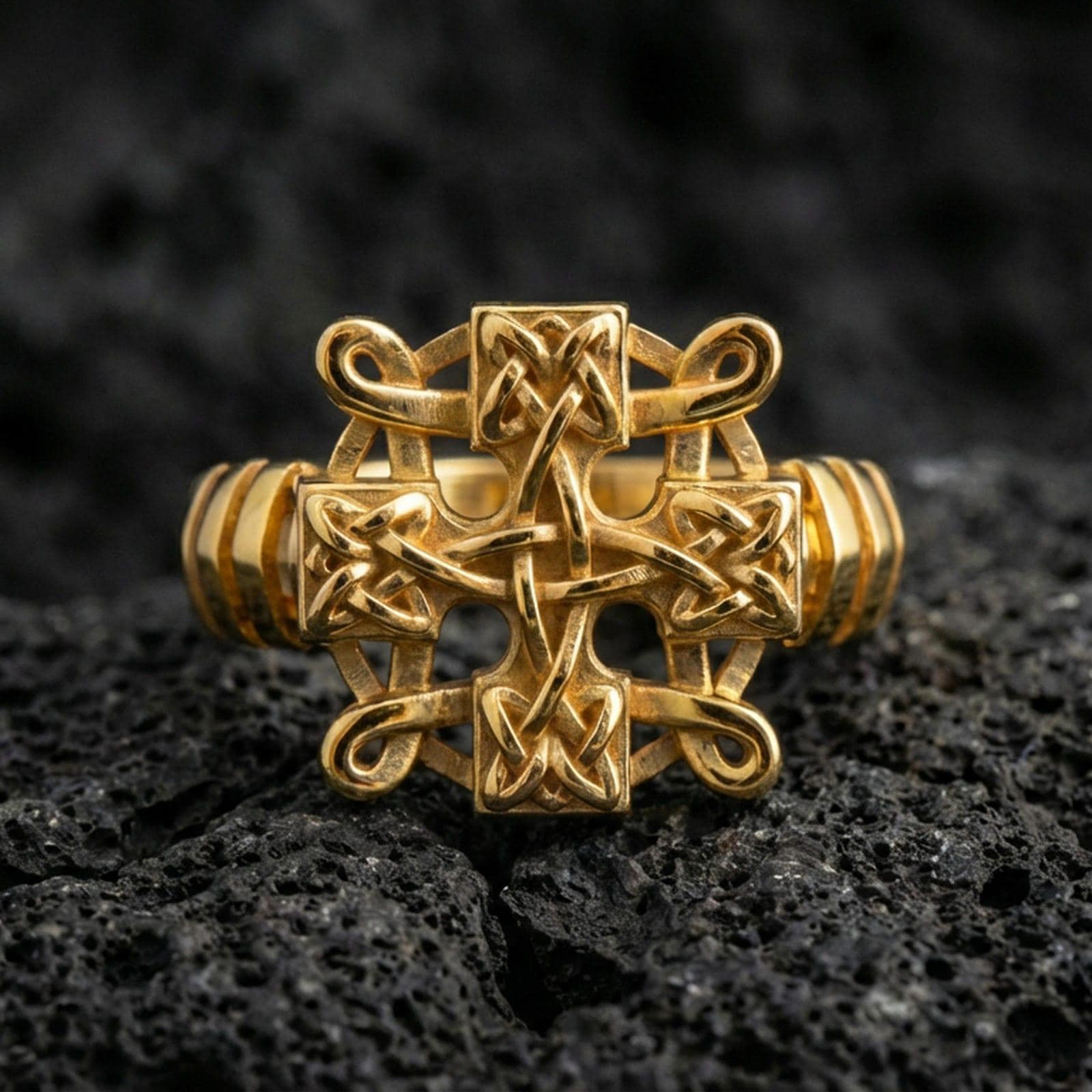 Gold plated Celtic knot cross ring with raised braided knot detailing and high-contrast relief, product-only macro image on black volcanic rock