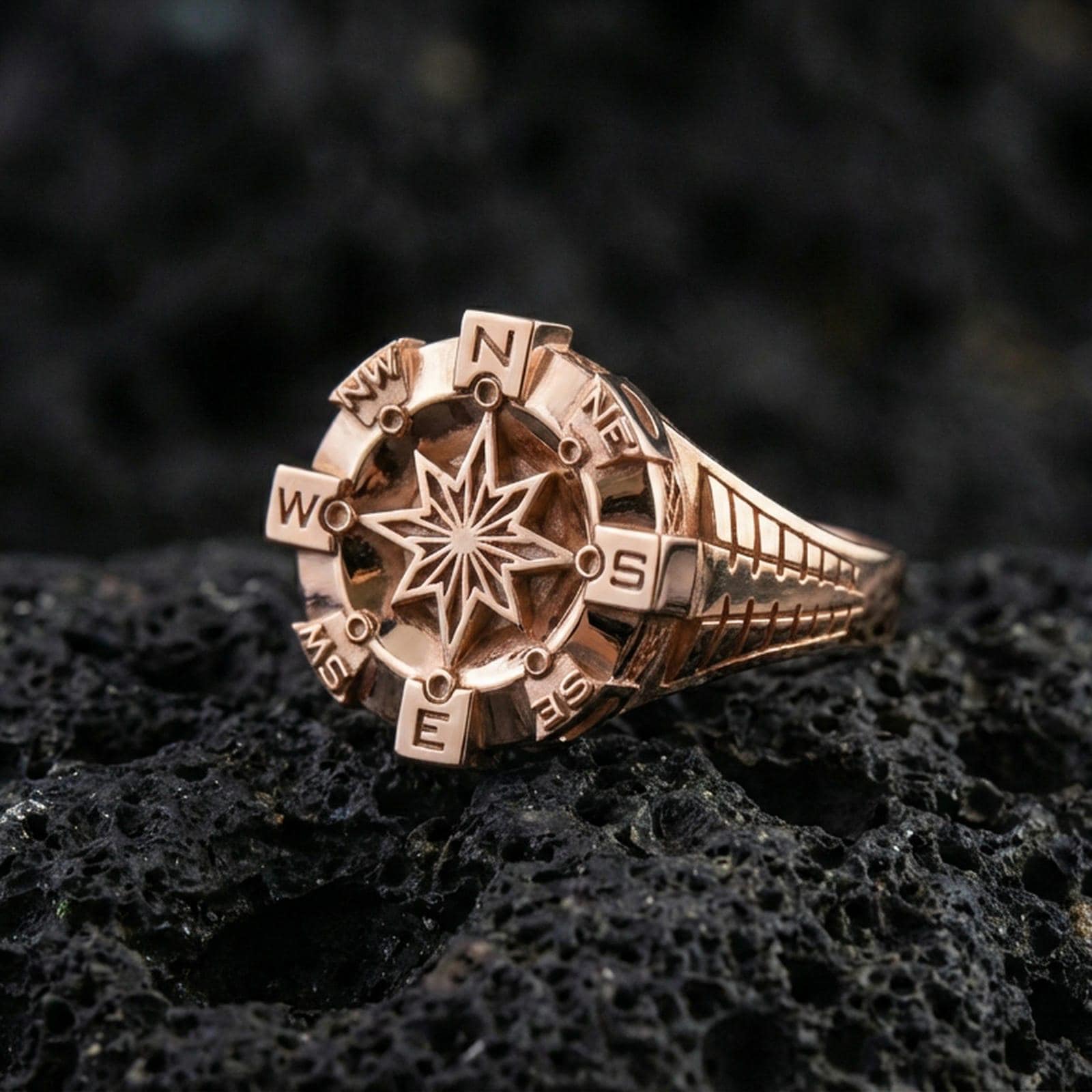Rose gold plated compass ring with sculpted navigation star and cardinal points, close-up product shot on volcanic rock