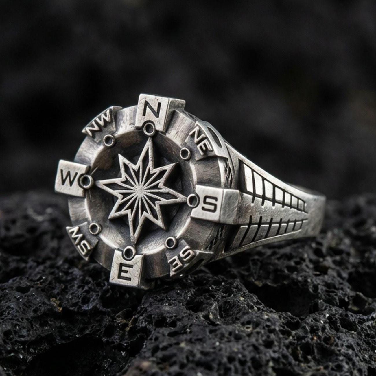 Oxidized sterling silver compass ring with raised eight-point star and cardinal direction markers, macro product photo on black volcanic rock