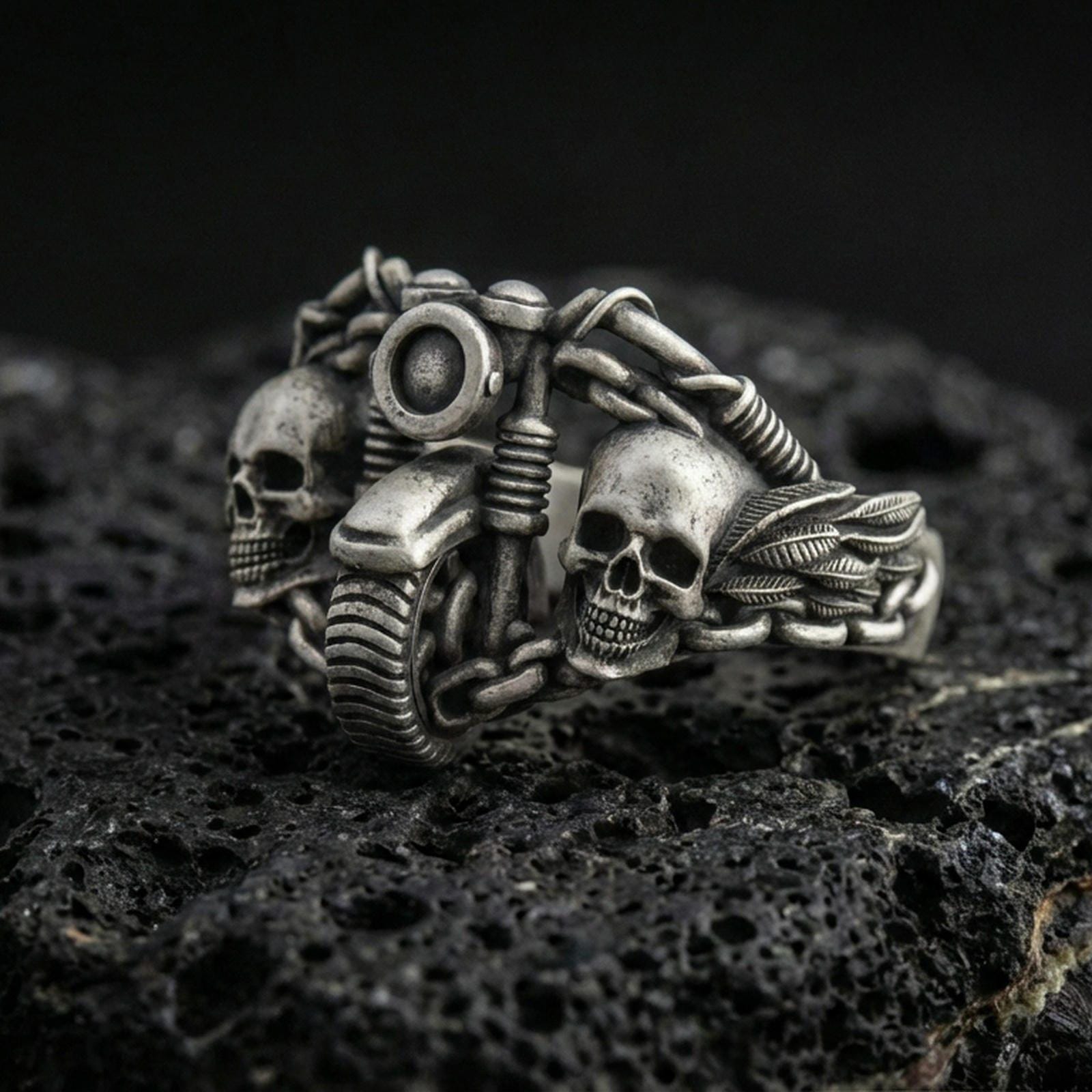 Oxidized sterling silver double skull motorcycle ring with detailed handlebars and chain links, macro product photo on black volcanic rock