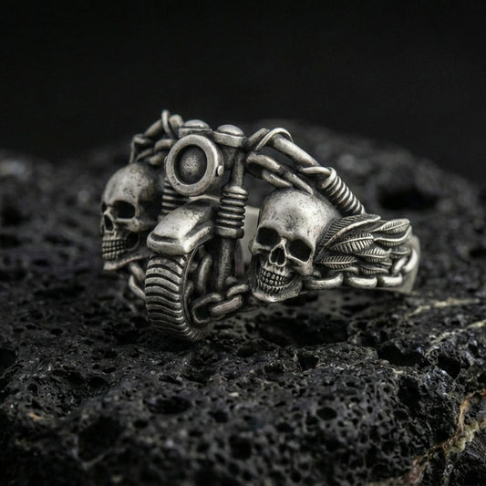Oxidized sterling silver double skull motorcycle ring with detailed handlebars and chain links, macro product photo on black volcanic rock