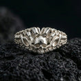Load image into Gallery viewer, Polished silver skull ring featuring raised spiked crest and industrial chain textures on the band, close-up on volcanic stone
