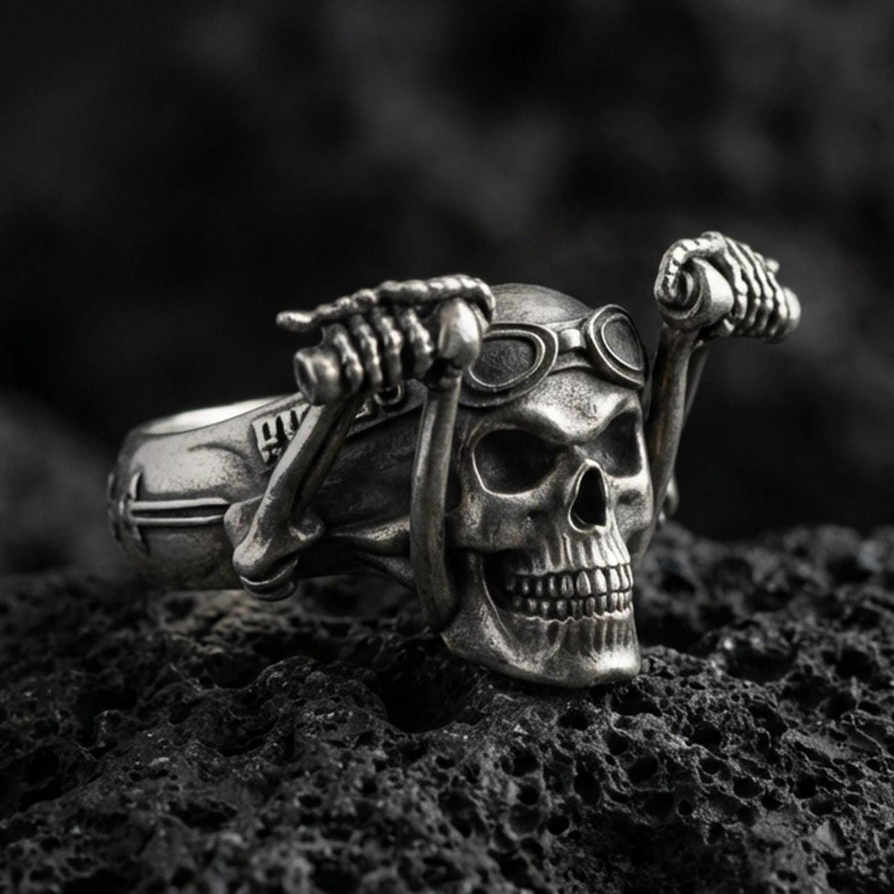 Oxidized sterling silver biker skull ring with aviator goggles and raised skeletal hands, macro product photo on black volcanic rock