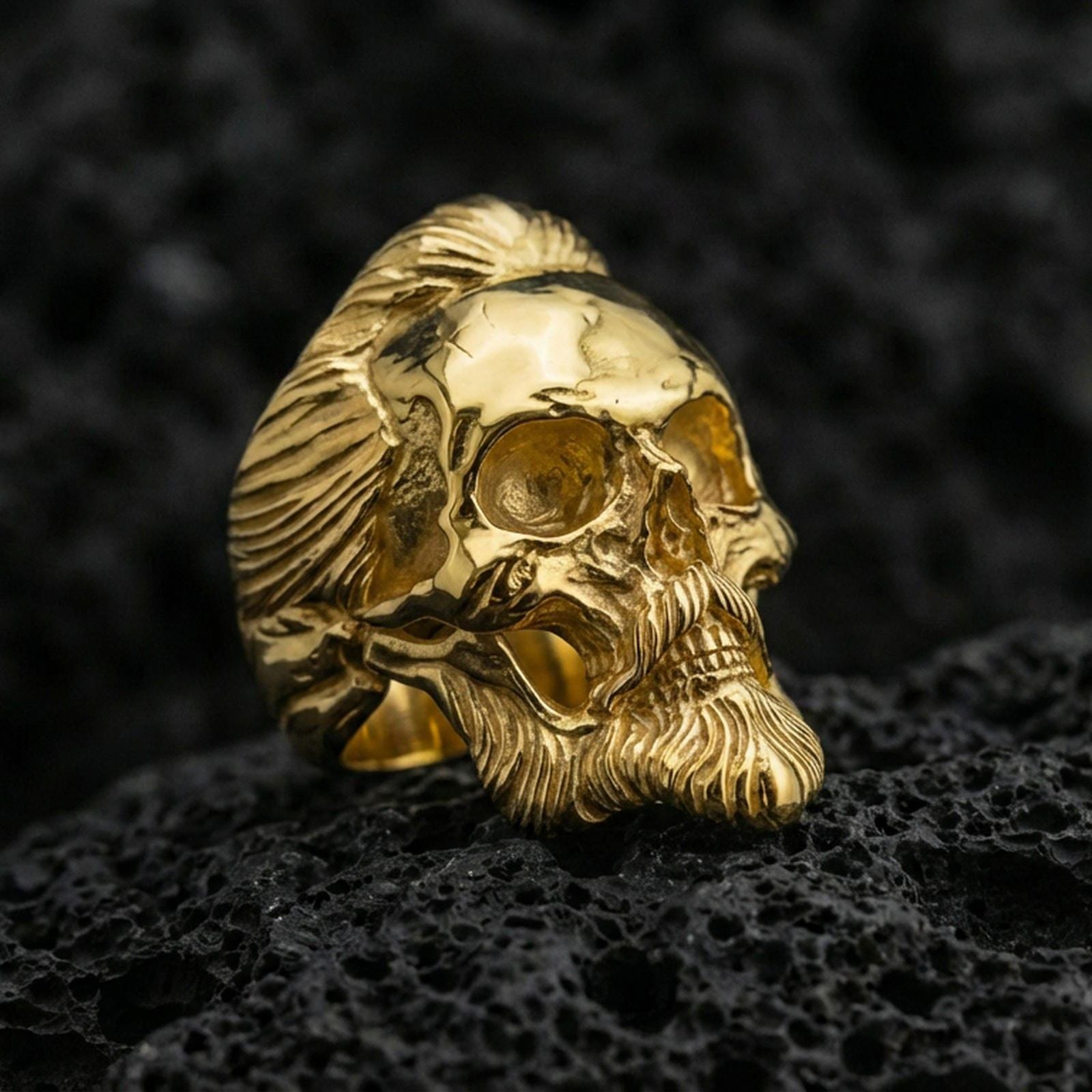 Gold plated skull ring featuring a carved beard and slicked-back hair, product-only macro image on black volcanic rock