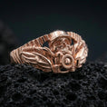 Load image into Gallery viewer, Rose gold plated winged skull biker ring with motorcycle-inspired details and rugged relief, close-up on volcanic rock
