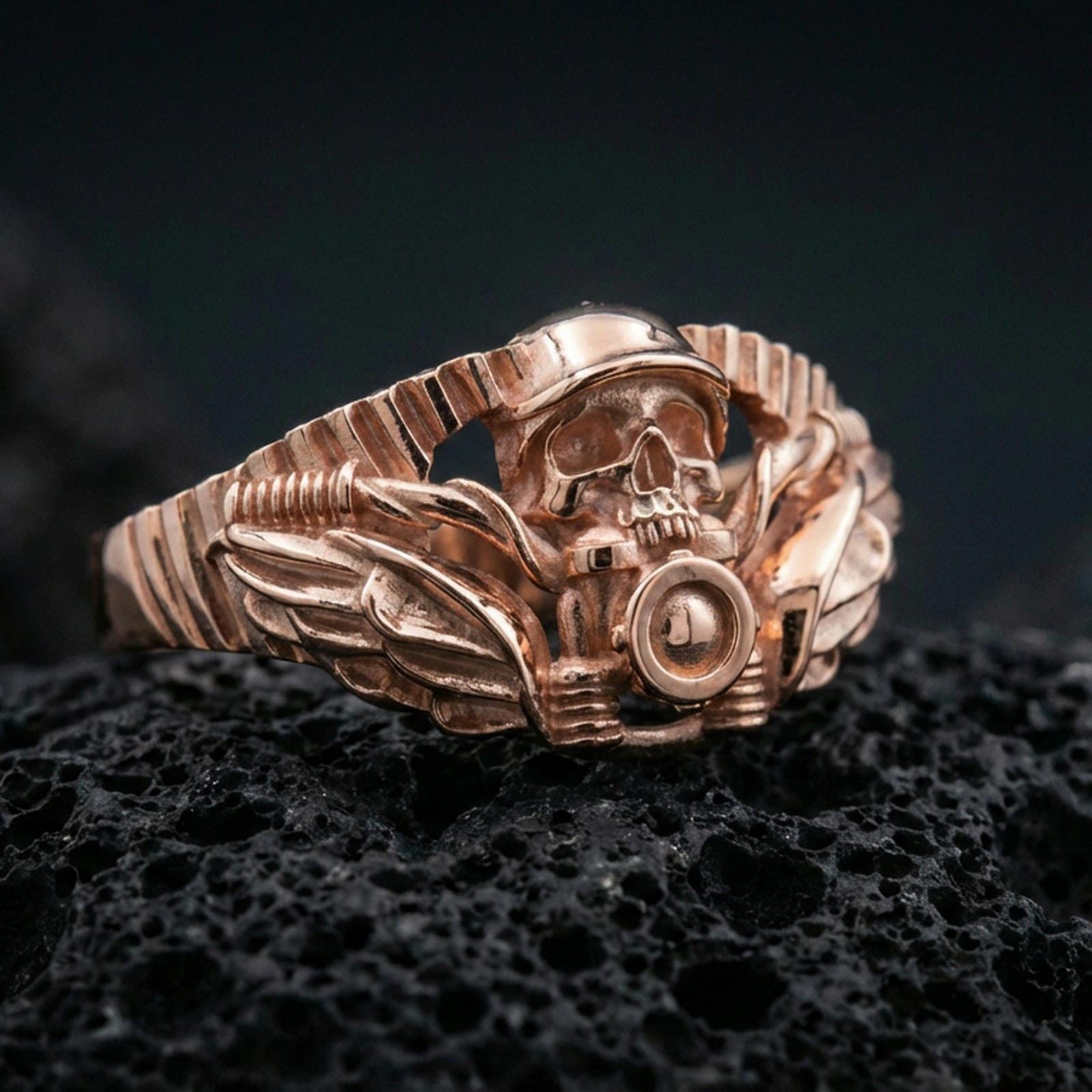 Rose gold plated winged skull biker ring with motorcycle-inspired details and rugged relief, close-up on volcanic rock