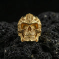 Load image into Gallery viewer, Gold plated biomechanical skull ring with sci-fi engraved patterning and bold skull face design, product-only macro on black volcanic rock
