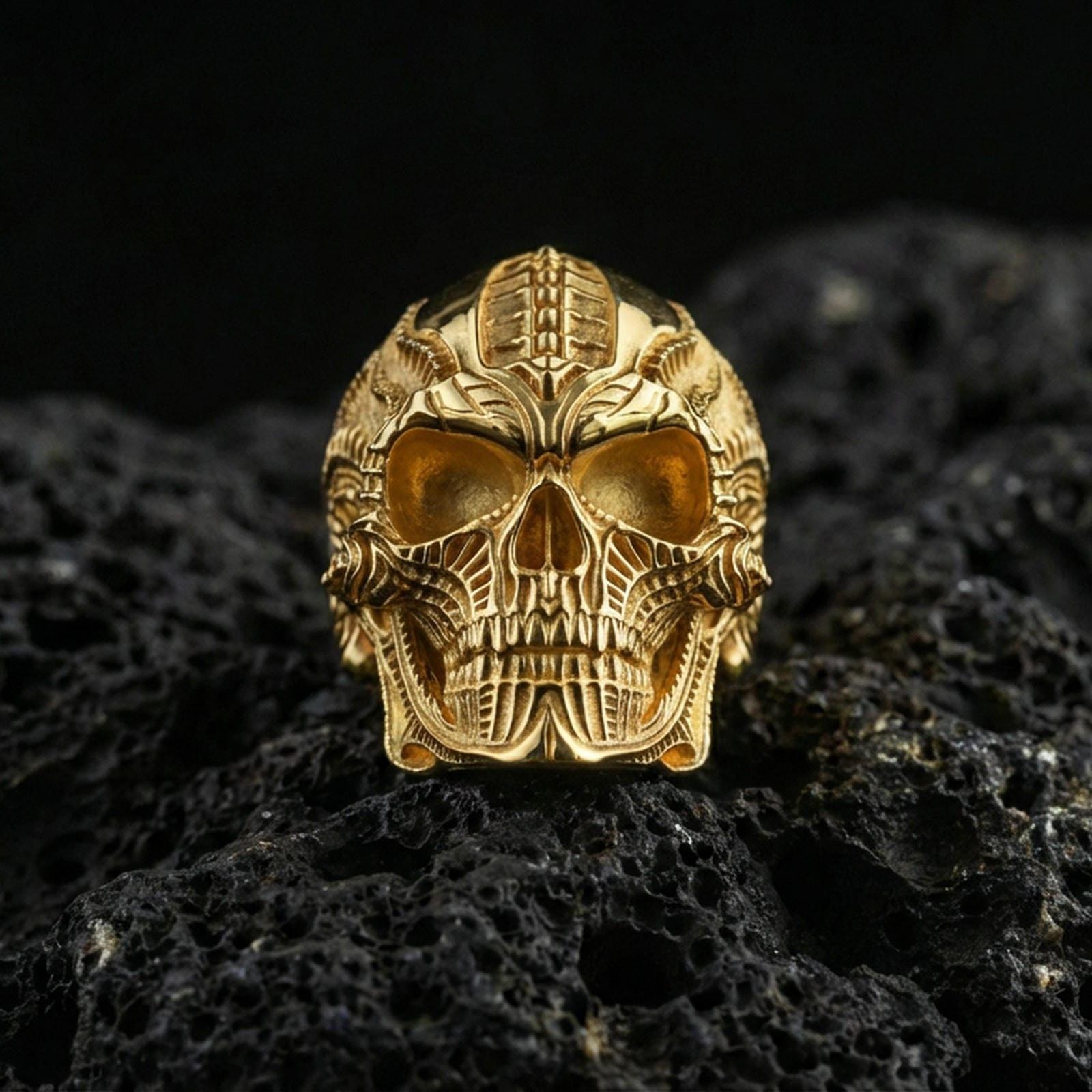 Gold plated biomechanical skull ring with sci-fi engraved patterning and bold skull face design, product-only macro on black volcanic rock