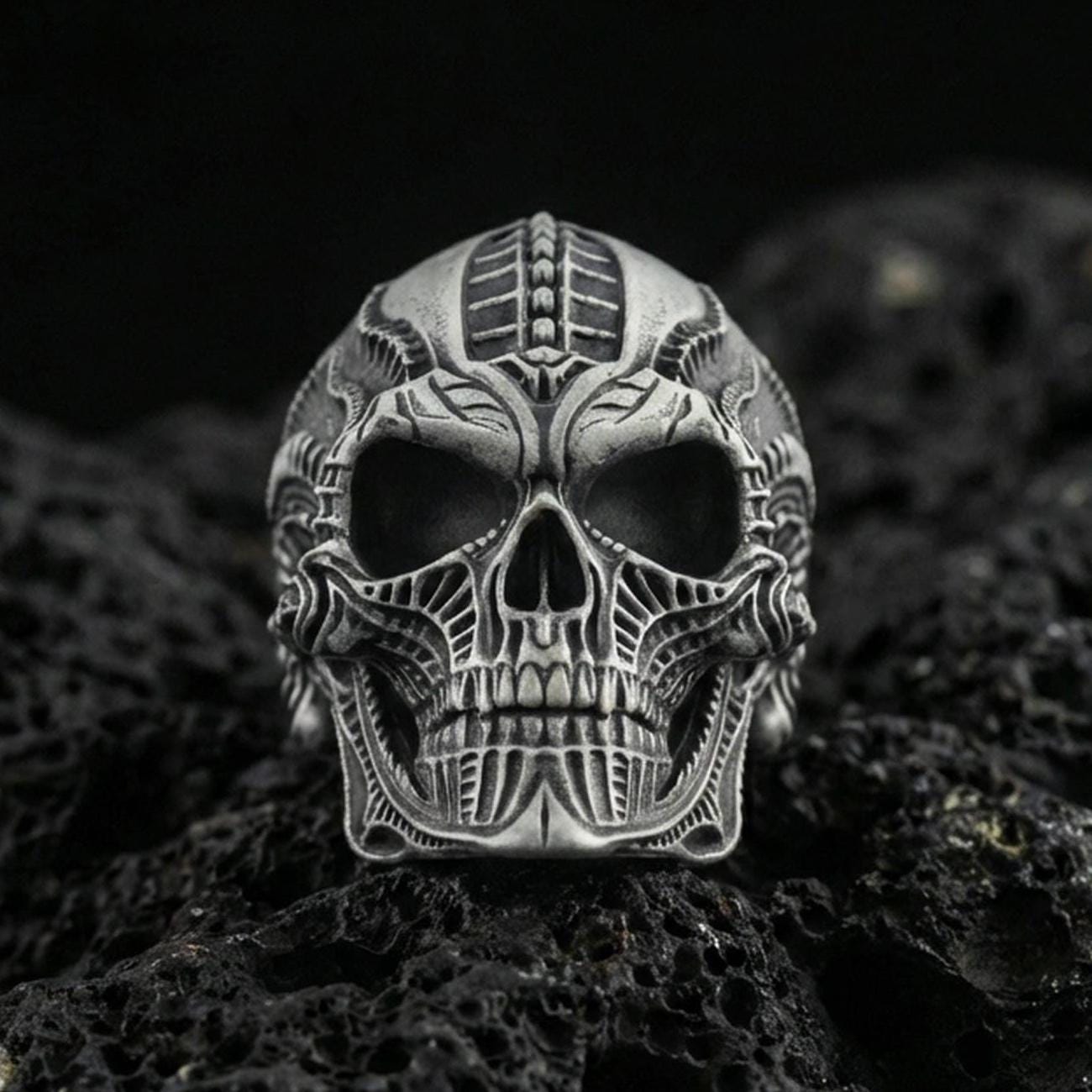 Oxidized sterling silver biomechanical skull ring with futuristic engraved lines and deep relief, macro photo on black volcanic rock