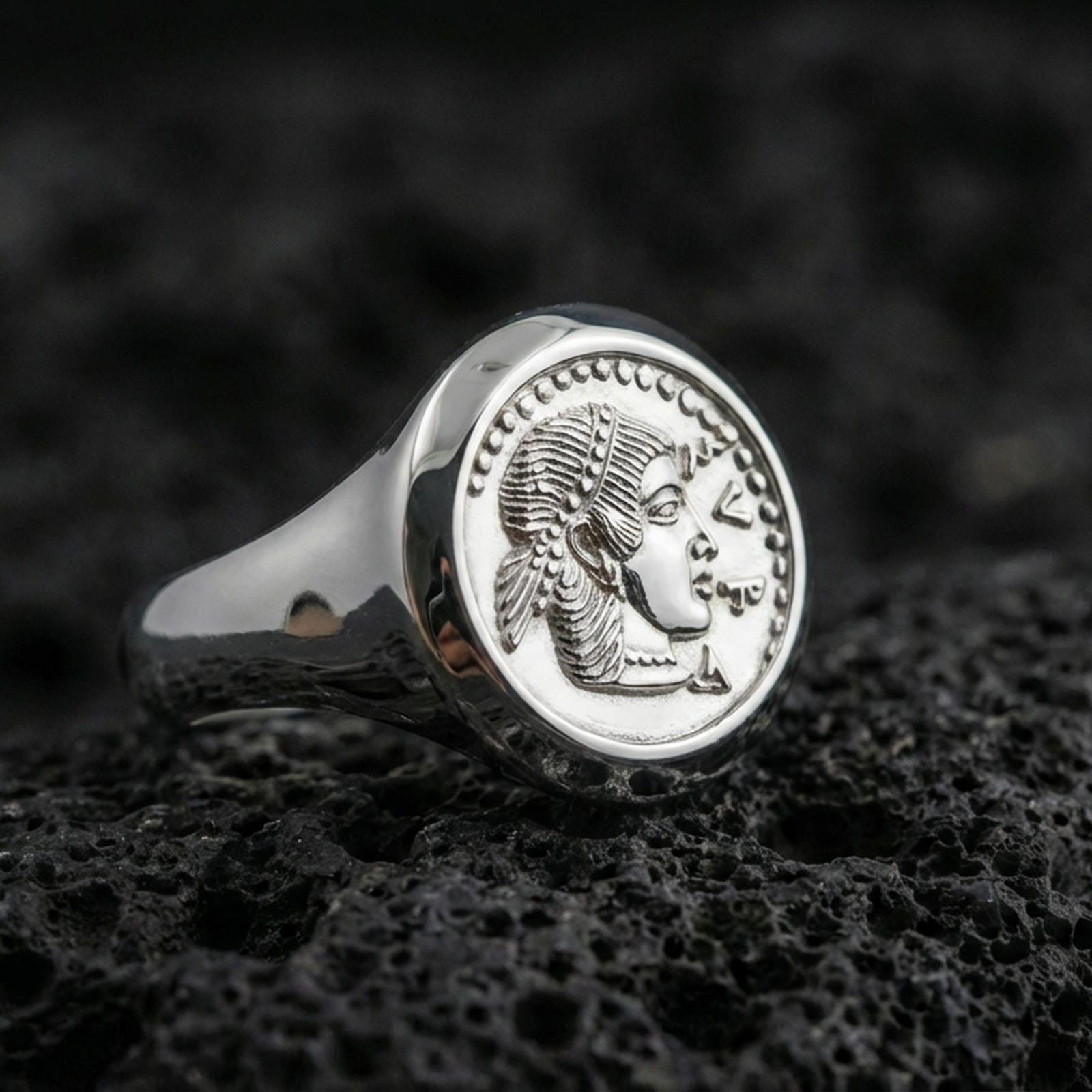 Polished silver Greek coin ring featuring a raised portrait relief and smooth signet band, close-up on volcanic stone