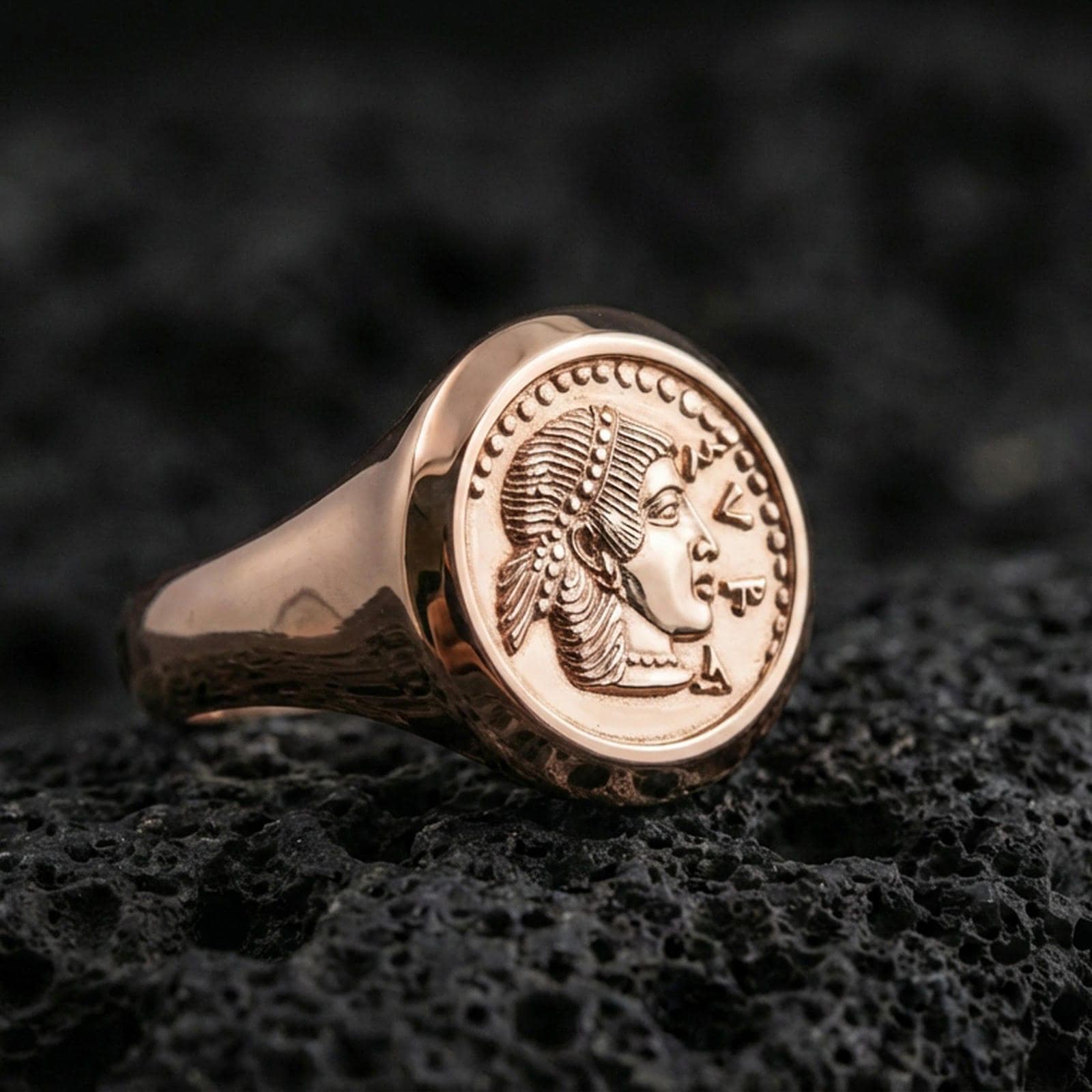 Rose gold plated Greek coin ring showing a vintage portrait coin face and rounded signet shank, close-up on volcanic rock