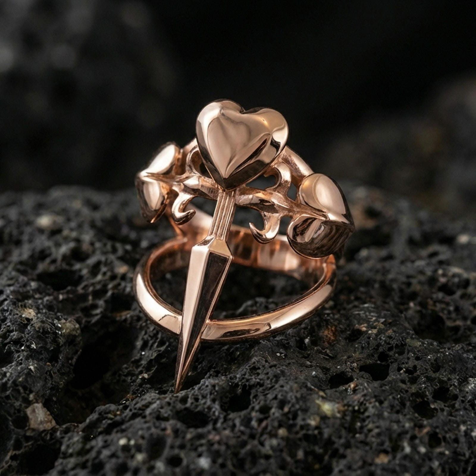 Rose gold plated heart dagger ring showing three hearts and an elongated dagger point, close-up on volcanic rock