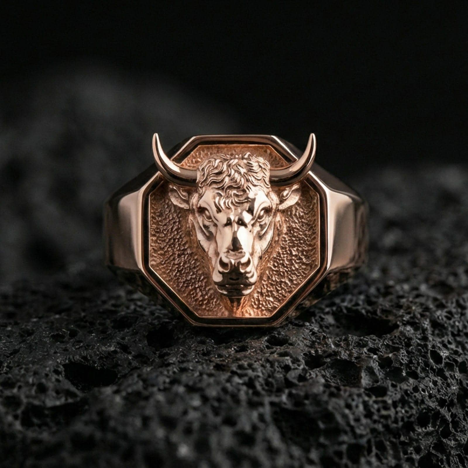 Rose gold plated bull head signet ring on volcanic rock, textured lava stone background, bold horns and sculpted face detail