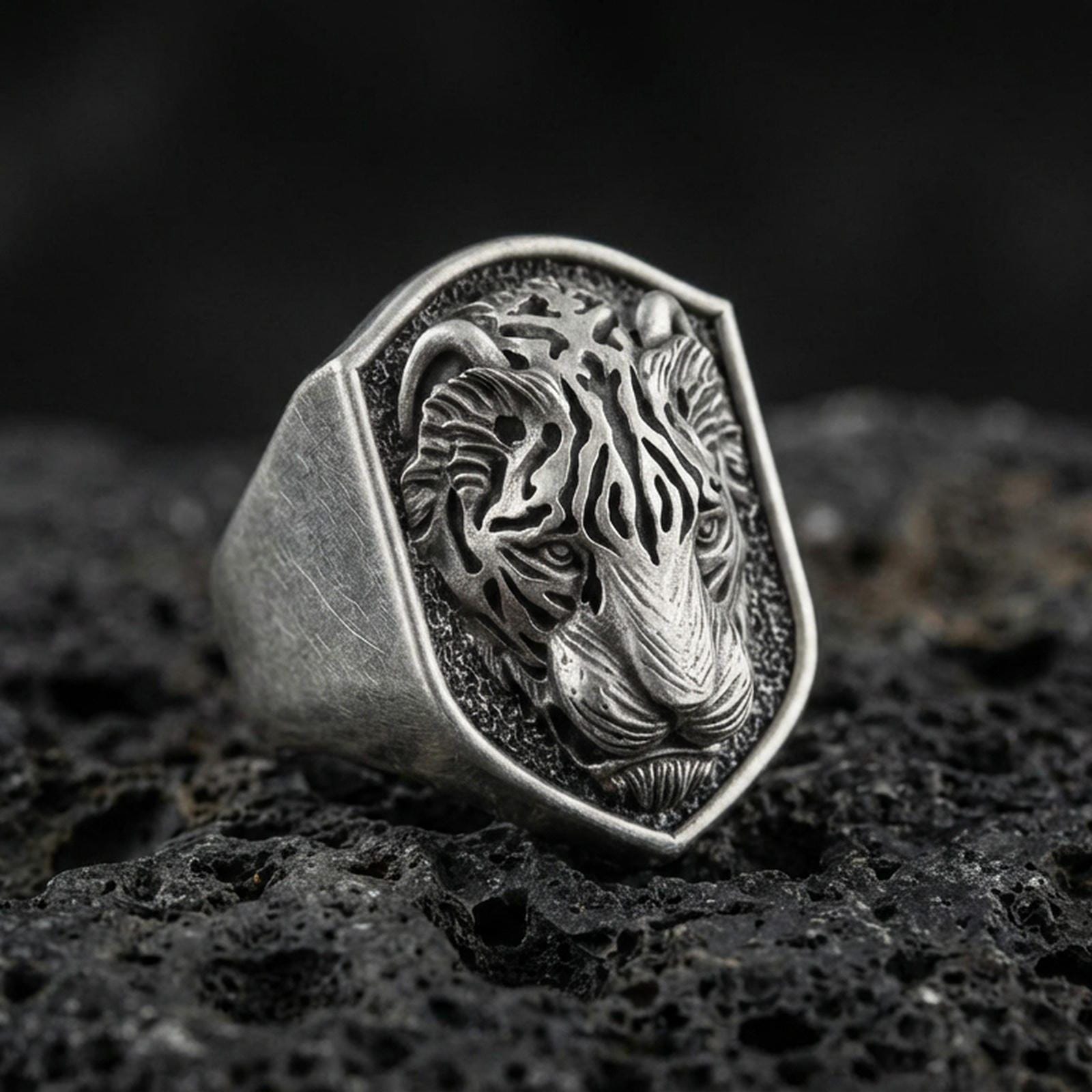 Oxidized sterling silver tiger signet ring on volcanic rock, shield-shaped face with raised tiger head relief and dark textured background, macro jewelry photo