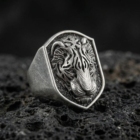 Oxidized sterling silver tiger signet ring on volcanic rock, shield-shaped face with raised tiger head relief and dark textured background, macro jewelry photo