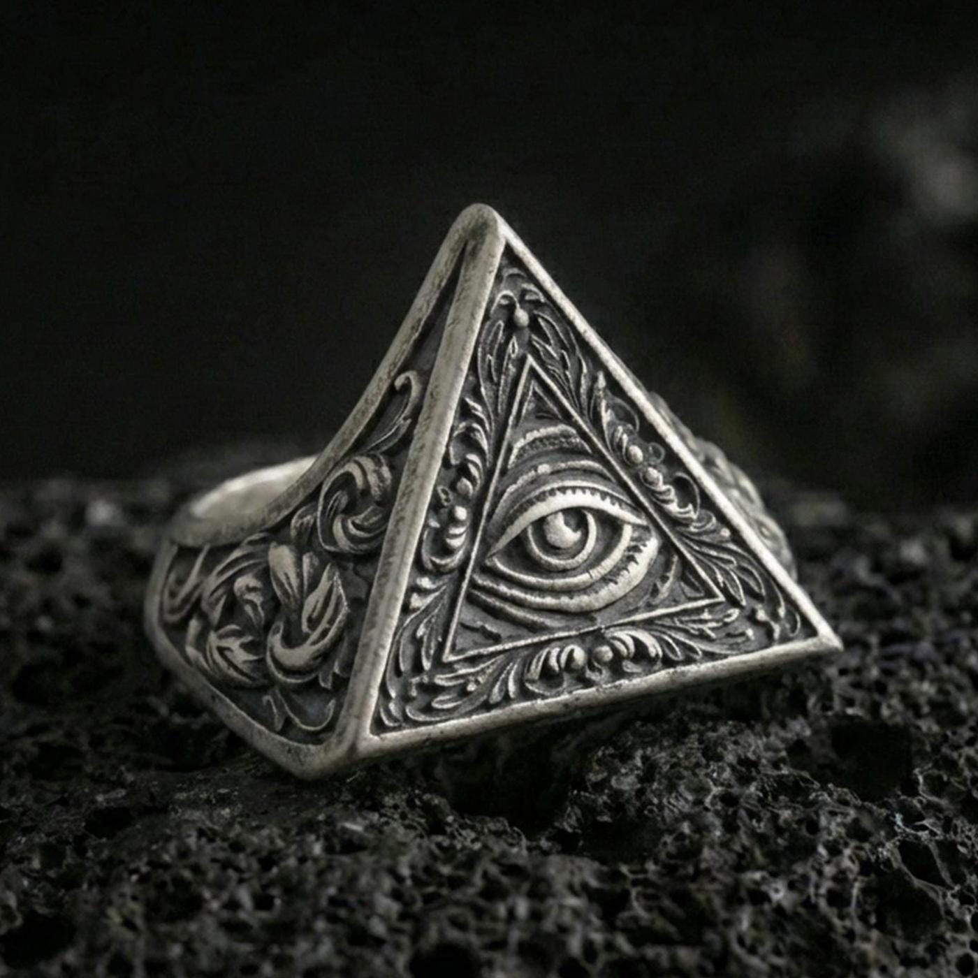Oxidized sterling silver triangle signet ring with all-seeing eye relief and ornate scroll engravings, macro shot on black volcanic rock
