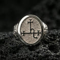 Load image into Gallery viewer, Polished silver oval signet ring featuring a carved occult sigil emblem on a grain-textured face, close-up on volcanic stone
