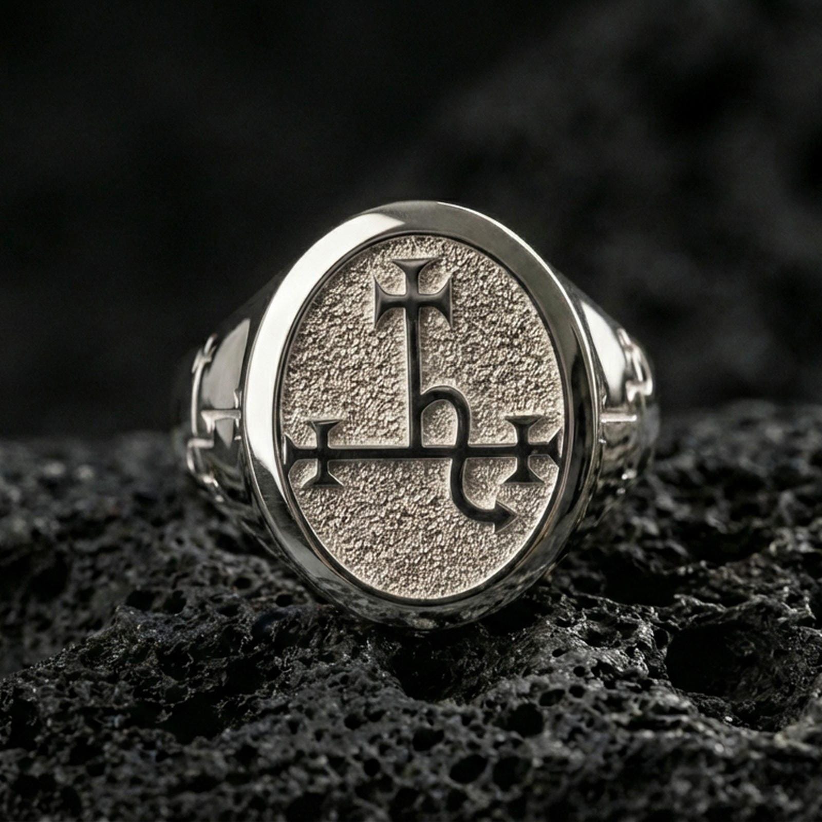 Polished silver oval signet ring featuring a carved occult sigil emblem on a grain-textured face, close-up on volcanic stone