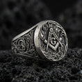 Load image into Gallery viewer, Oxidized sterling silver Masonic signet ring with square and compass “G” emblem and ornate scrollwork, on black volcanic rock
