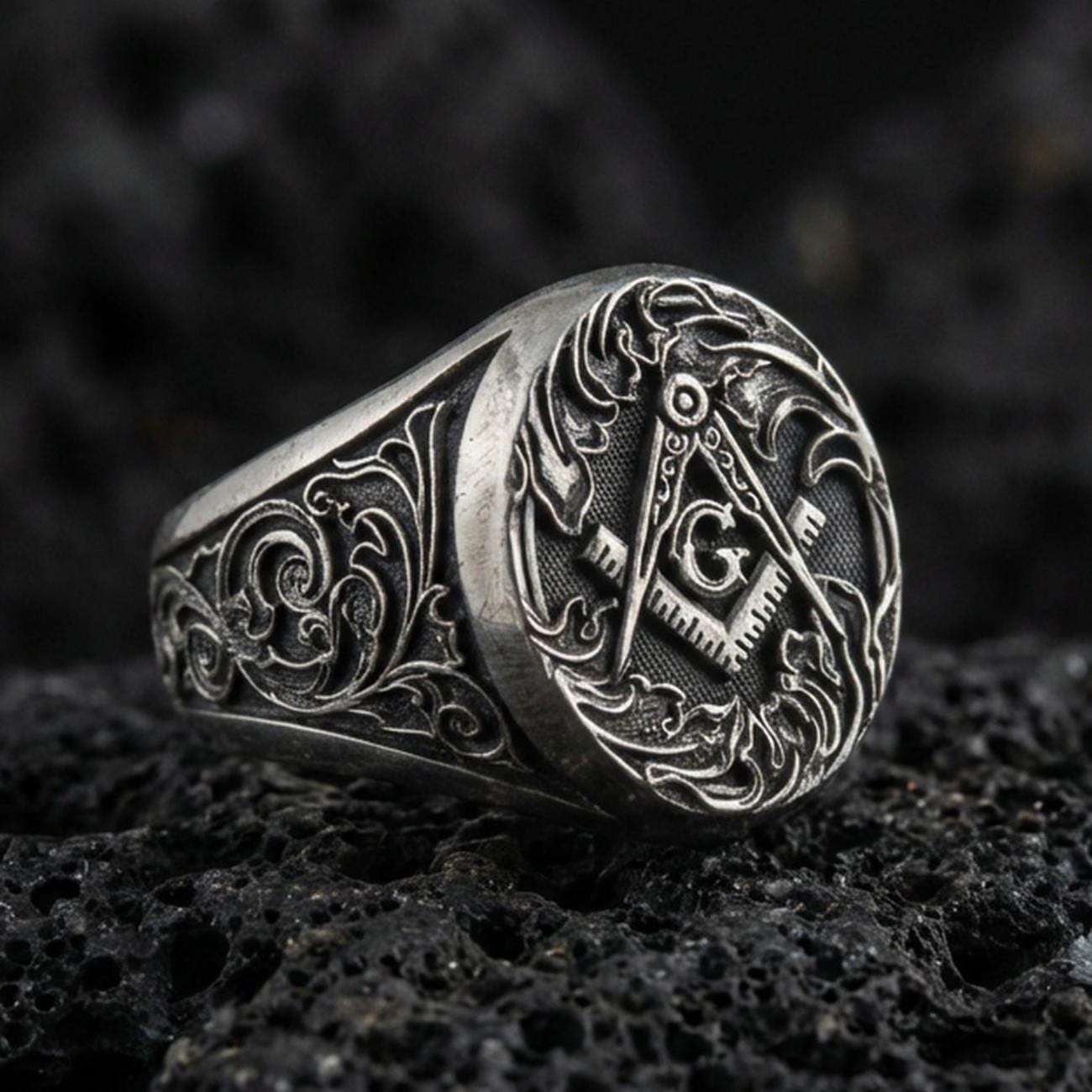 Oxidized sterling silver Masonic signet ring with square and compass “G” emblem and ornate scrollwork, on black volcanic rock