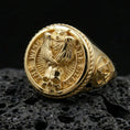 Load image into Gallery viewer, Gold plated owl and skull signet ring with circular quote engraving and braided edge frame, close-up on volcanic rock background
