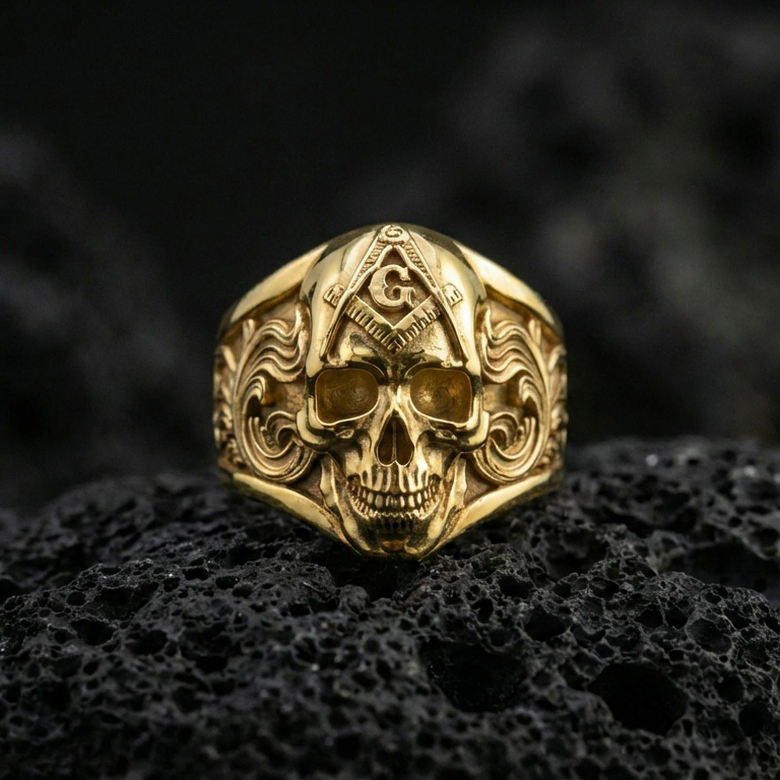 Gold plated Masonic skull ring with compass-and-G symbol and detailed baroque scroll patterns, close-up on volcanic rock background