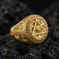 Load image into Gallery viewer, Gold plated all-seeing eye pyramid ring with detailed scrollwork band and Latin text around the face, close-up on volcanic rock
