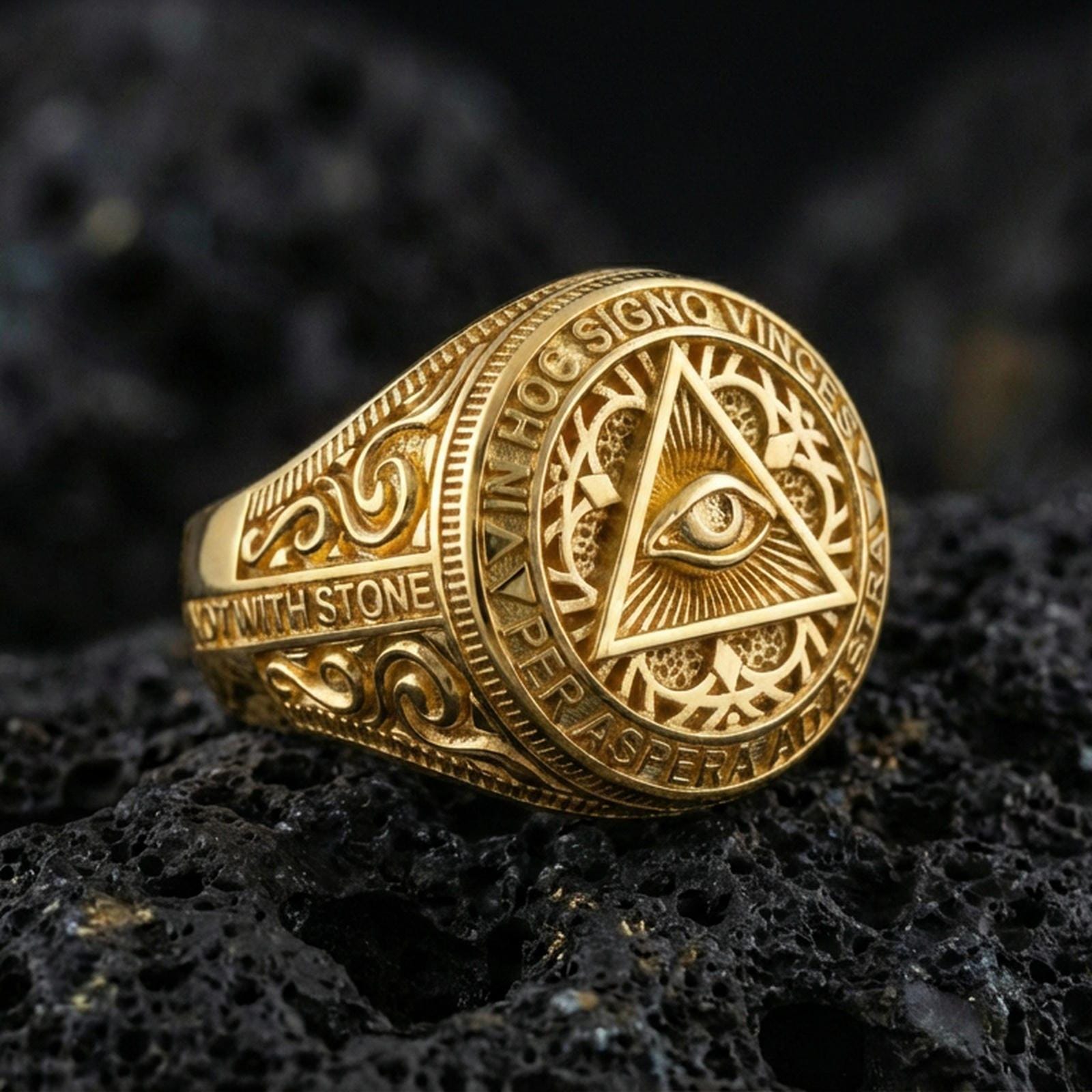 Gold plated all-seeing eye pyramid ring with detailed scrollwork band and Latin text around the face, close-up on volcanic rock