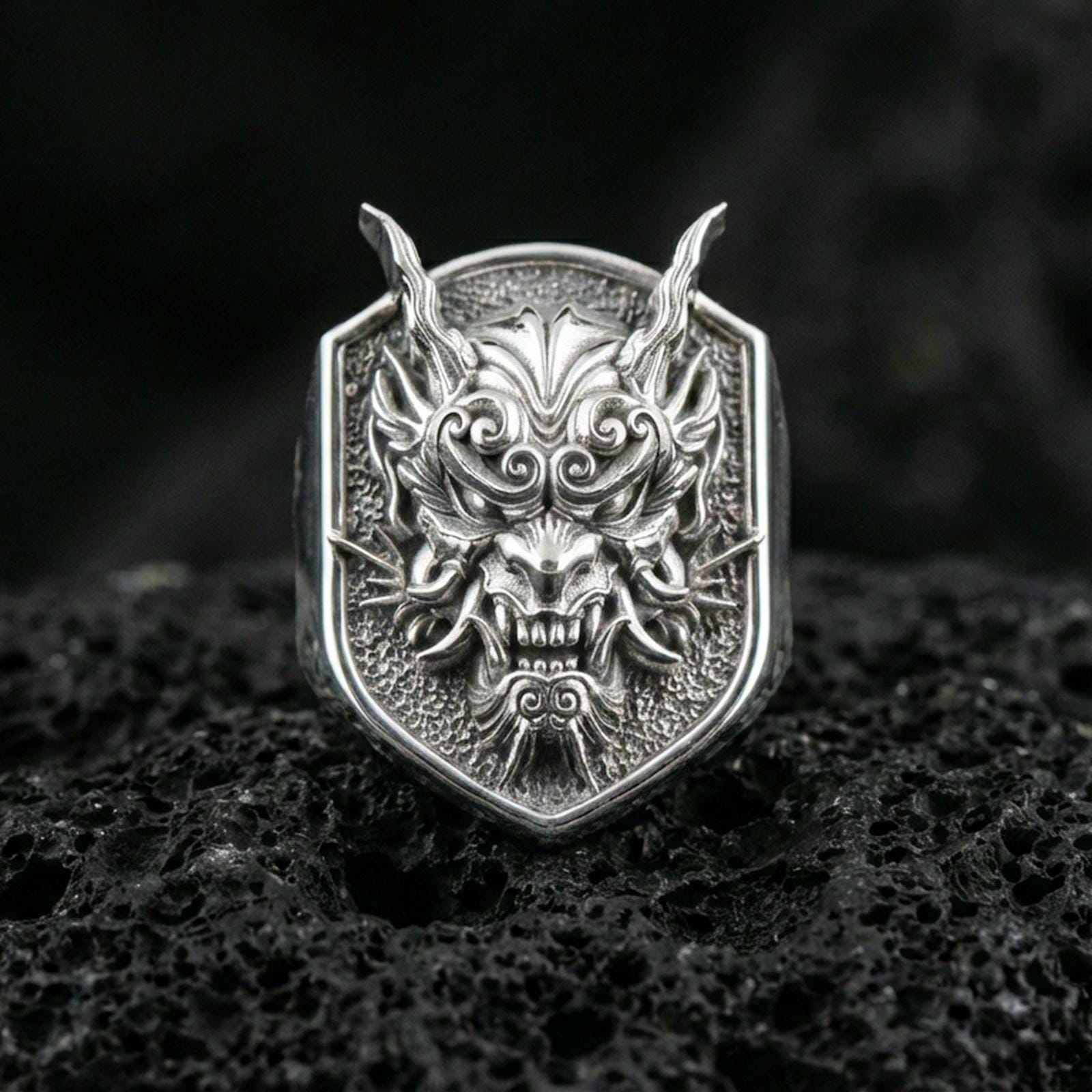 Polished silver Japanese oni mask ring with sharp horns and ornate swirl detailing, close-up on dark volcanic stone background