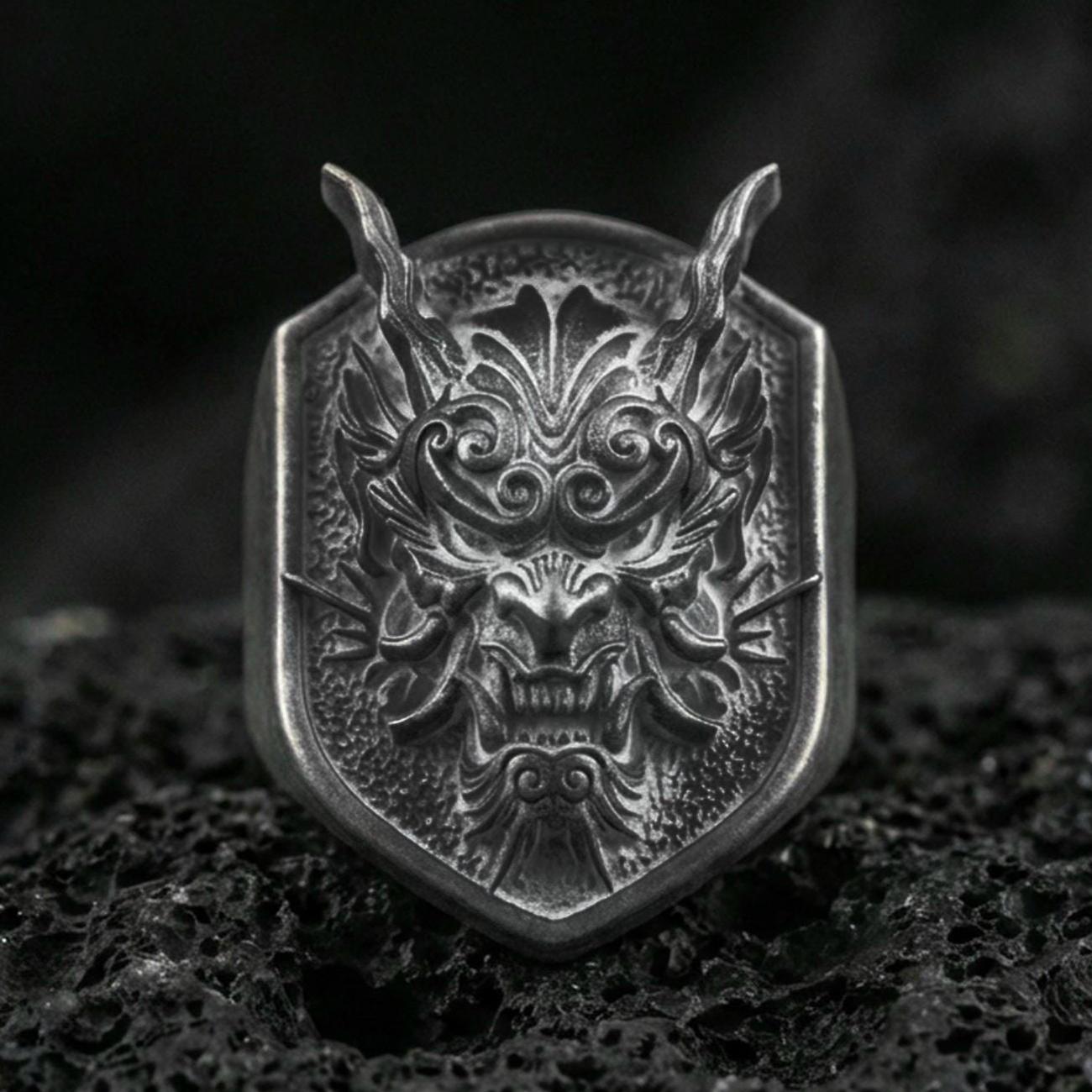 Oxidized sterling silver oni mask signet ring with horned Japanese demon face and deep relief texture, macro on black volcanic rock